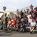 A group of U.S. Marines in camouflage uniforms are addressing a large civilian audience seated on bleachers at what appears to be a military base. The civilians, many wearing lanyards and red shirts, appear to be attending an organized event such as a family day, community outreach, or recruit graduation. Palm trees and military-style buildings are visible in the background, suggesting a Southern California installation.
