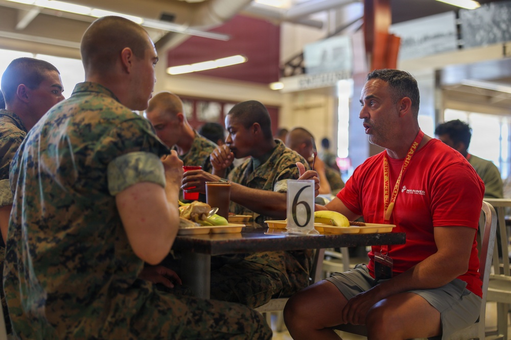 A group of individuals in U.S. Marine Corps camouflage uniforms are seated at a cafeteria-style dining table eating a meal. A civilian or recruiter in a red 'Marines' branded shirt and lanyard is engaged in conversation with one of the uniformed personnel. The setting appears to be a military mess hall or dining facility, with a table number marker '6' visible.