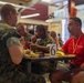 A group of individuals in U.S. Marine Corps camouflage uniforms are seated at a cafeteria-style dining table eating a meal. A civilian or recruiter in a red 'Marines' branded shirt and lanyard is engaged in conversation with one of the uniformed personnel. The setting appears to be a military mess hall or dining facility, with a table number marker '6' visible.