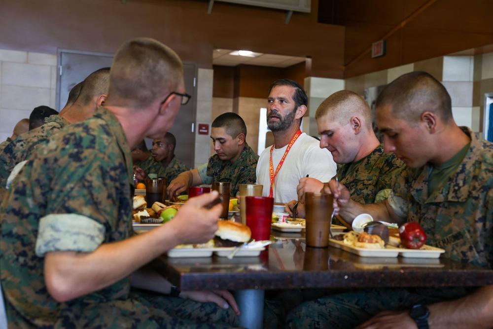 A group of military personnel in camouflage uniforms are seated at a dining table in what appears to be a military mess hall or chow hall. One civilian man wearing a white t-shirt and an orange lanyard/badge is seated among them, appearing to be a visitor or guest. The group is eating a meal together with various food items and drinks visible on the table.