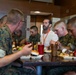 A group of military personnel in camouflage uniforms are seated at a dining table in what appears to be a military mess hall or chow hall. One civilian man wearing a white t-shirt and an orange lanyard/badge is seated among them, appearing to be a visitor or guest. The group is eating a meal together with various food items and drinks visible on the table.