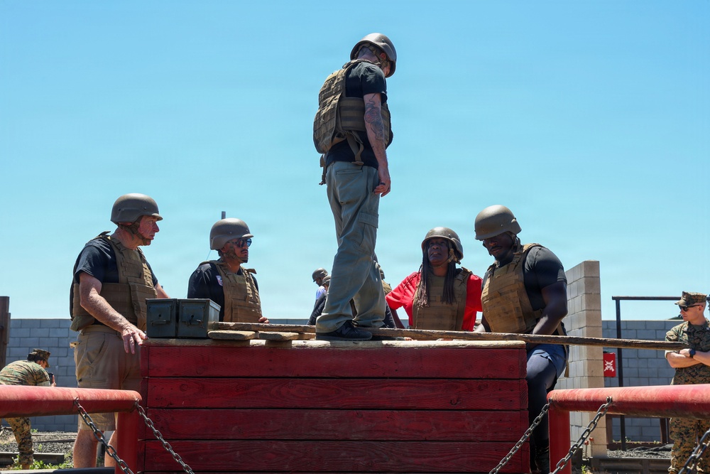 A group of civilians wearing tactical vests and helmets are standing on top of a red wooden obstacle structure, likely at a military training facility. They appear to be participating in a supervised experiential or media event alongside uniformed Marines visible in the background. The setting suggests a military base obstacle course or demonstration area.
