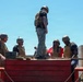 A group of civilians wearing tactical vests and helmets are standing on top of a red wooden obstacle structure, likely at a military training facility. They appear to be participating in a supervised experiential or media event alongside uniformed Marines visible in the background. The setting suggests a military base obstacle course or demonstration area.