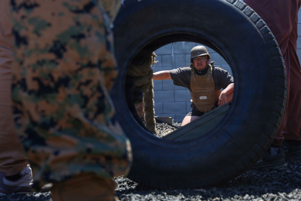 A person wearing a tactical vest and helmet is visible through the opening of a large tire, appearing to be engaged in a military or law enforcement training exercise. In the foreground, another individual in camouflage uniform is partially visible. The setting appears to be an outdoor training course with gravel ground and a concrete block wall in the background.