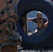 A person wearing a tactical vest and helmet is visible through the opening of a large tire, appearing to be engaged in a military or law enforcement training exercise. In the foreground, another individual in camouflage uniform is partially visible. The setting appears to be an outdoor training course with gravel ground and a concrete block wall in the background.