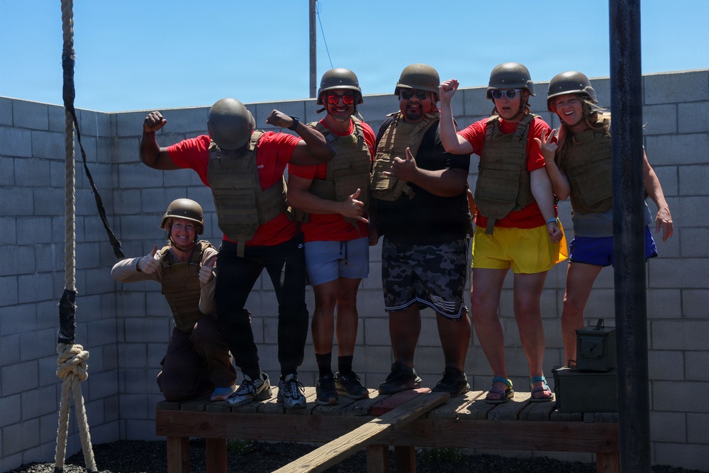 A group of six people pose enthusiastically at what appears to be a military-themed activity or experience venue. All participants are wearing olive/tan tactical vests and military-style helmets, with most wearing red shirts. They are standing on a wooden platform against a concrete block wall, striking celebratory poses including flexing and thumbs-up gestures.