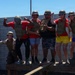 A group of six people pose enthusiastically at what appears to be a military-themed activity or experience venue. All participants are wearing olive/tan tactical vests and military-style helmets, with most wearing red shirts. They are standing on a wooden platform against a concrete block wall, striking celebratory poses including flexing and thumbs-up gestures.