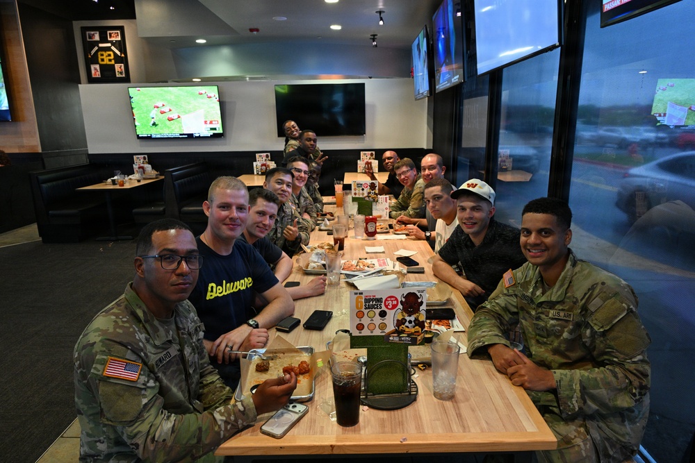 A group of approximately 12-14 military personnel, mostly in U.S. Army camouflage uniforms, are gathered at a restaurant that appears to be Buffalo Wild Wings, sharing a meal together. Several members are in civilian clothing mixed among those in uniform. The group is seated at joined tables and posing for a group photo in what appears to be a casual dining outing.
