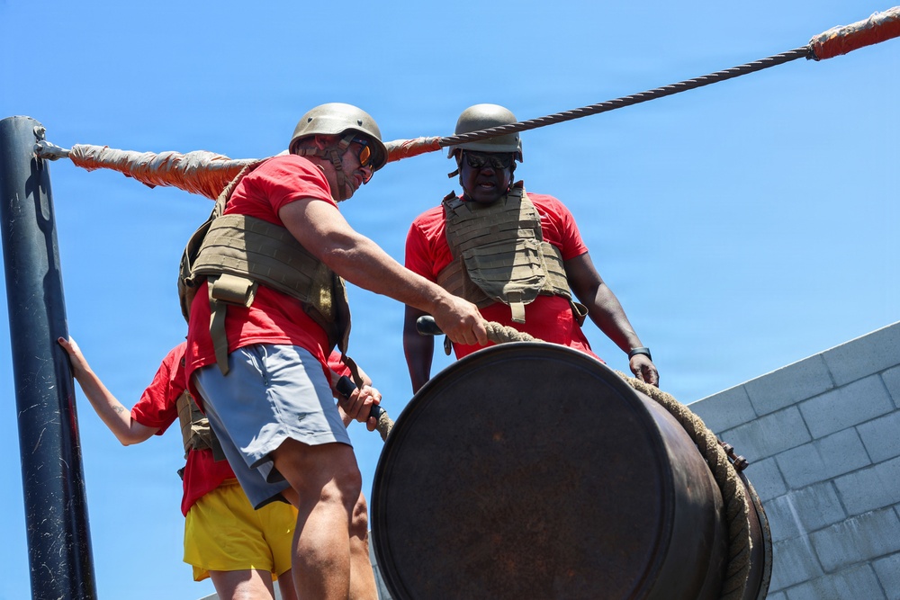 Two individuals wearing matching red t-shirts, tactical/plate carrier vests, and military-style helmets are working together to lift or maneuver a large heavy metal barrel using a rope. A third person in a red shirt and yellow shorts is partially visible in the background. The setting appears to be an outdoor obstacle course or physical training environment under bright sunny skies.