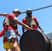 Two individuals wearing matching red t-shirts, tactical/plate carrier vests, and military-style helmets are working together to lift or maneuver a large heavy metal barrel using a rope. A third person in a red shirt and yellow shorts is partially visible in the background. The setting appears to be an outdoor obstacle course or physical training environment under bright sunny skies.