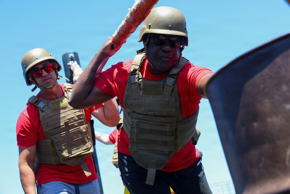 Two individuals wearing matching red t-shirts, tactical/plate carrier vests, ballistic helmets, and sunglasses are engaged in what appears to be a physical training or combat drill exercise. The foreground figure is actively swinging or wielding a padded orange pole/staff, suggesting a combatives or close-quarters training scenario. The setting appears to be outdoors under bright, clear blue sky conditions.