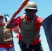 Two individuals wearing matching red t-shirts, tactical/plate carrier vests, ballistic helmets, and sunglasses are engaged in what appears to be a physical training or combat drill exercise. The foreground figure is actively swinging or wielding a padded orange pole/staff, suggesting a combatives or close-quarters training scenario. The setting appears to be outdoors under bright, clear blue sky conditions.