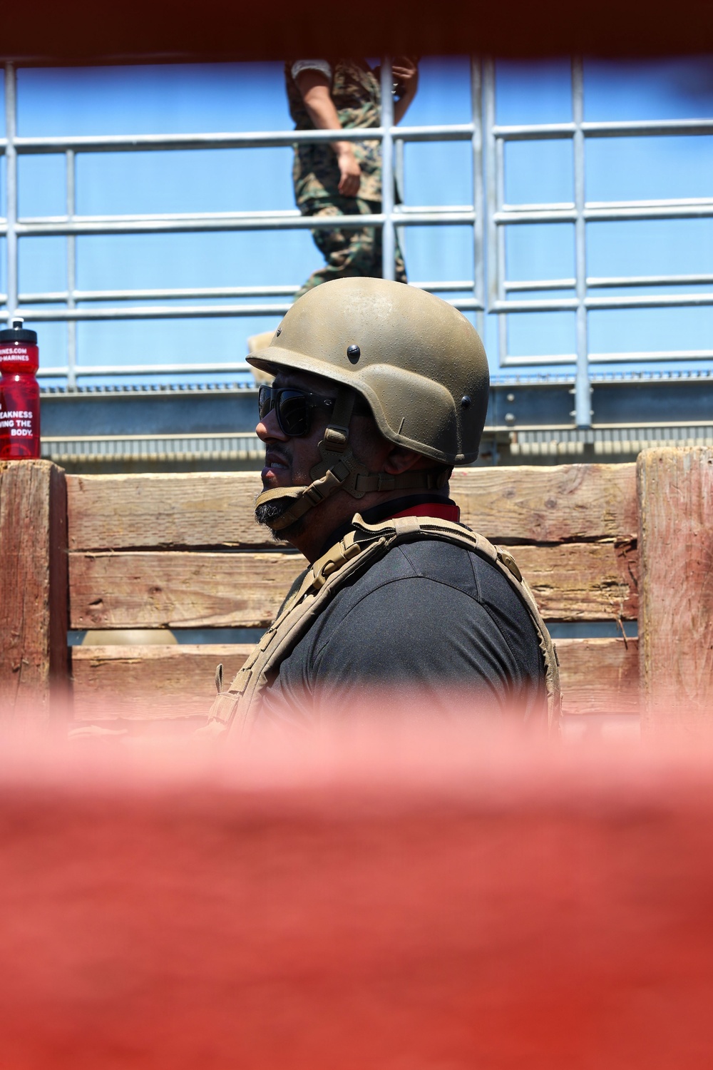 A person wearing a tan/olive military-style helmet, tactical vest, and dark sunglasses is visible in profile view, positioned behind a wooden barrier. In the background, a second individual in camouflage uniform is visible on an elevated platform or bleachers. A red water bottle with partial text referencing 'Marines' is visible to the left.