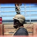 A person wearing a tan/olive military-style helmet, tactical vest, and dark sunglasses is visible in profile view, positioned behind a wooden barrier. In the background, a second individual in camouflage uniform is visible on an elevated platform or bleachers. A red water bottle with partial text referencing 'Marines' is visible to the left.
