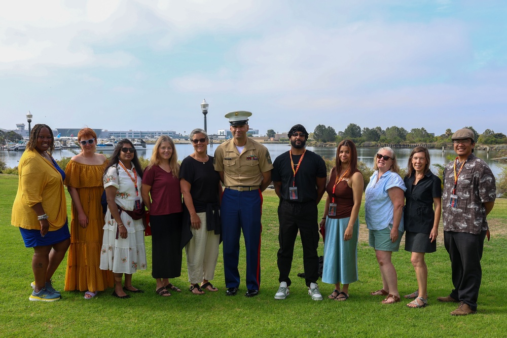 A group of eleven people pose for a group photo on a grassy area near a waterfront or marina. One individual in the center is wearing a United States Marine Corps dress uniform, while the others appear to be civilians, several of whom are wearing red lanyards with ID badges. The background features a calm body of water, trees, and what appears to be an airport or industrial facility in the distance.
