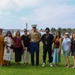 A group of eleven people pose for a group photo on a grassy area near a waterfront or marina. One individual in the center is wearing a United States Marine Corps dress uniform, while the others appear to be civilians, several of whom are wearing red lanyards with ID badges. The background features a calm body of water, trees, and what appears to be an airport or industrial facility in the distance.
