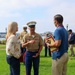 A U.S. Marine in dress uniform is engaged in conversation with two civilians on a grassy outdoor area near a waterfront. The Marine appears to be speaking with a woman in a floral blouse and a man in a blue polo shirt who is holding printed materials. Additional Marines and civilians are visible in the background.