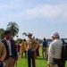 An outdoor gathering on a grassy area near a waterfront, featuring at least two U.S. Marine Corps personnel in Service 'C' (khaki) uniforms with blue trousers. Civilians and military members appear to be mingling or attending a public event, possibly a community outreach or military appreciation event. Palm trees and a body of water are visible in the background, suggesting a Southern California coastal location.