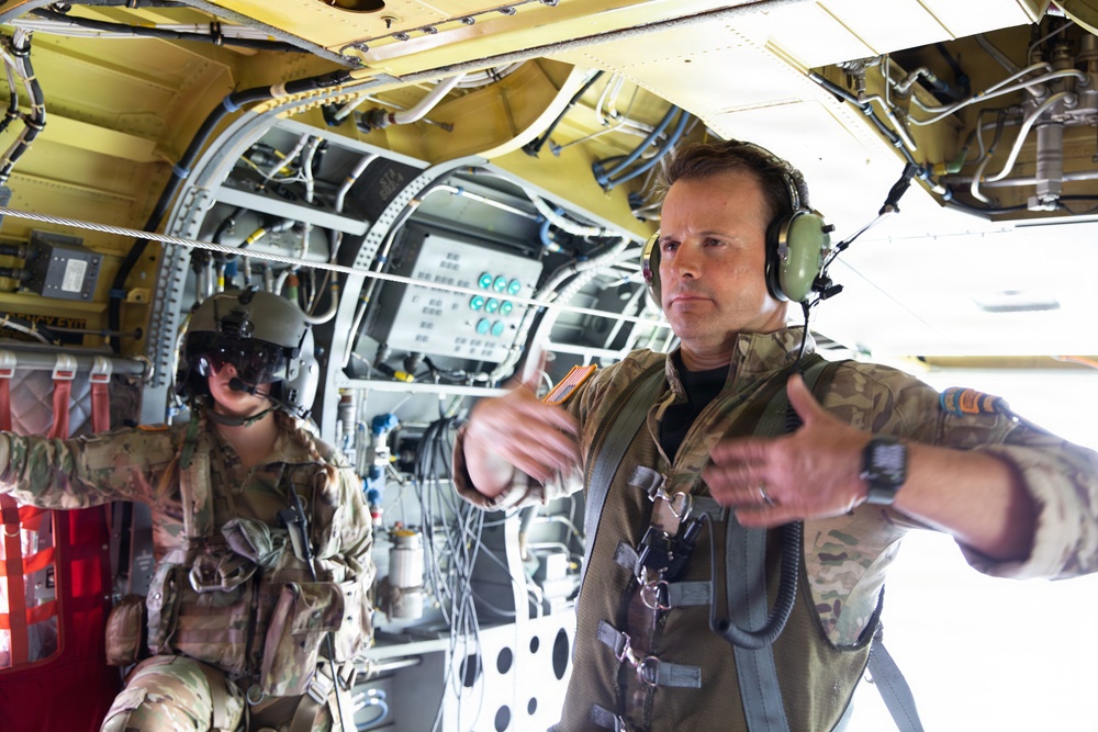 Two military personnel are photographed inside the cargo/crew compartment of a large military aircraft, likely a CH-47 Chinook or similar heavy-lift helicopter. The foreground figure, wearing a flight suit, multicam uniform, and aviation headset, appears to be gesturing with both arms extended outward. The interior shows exposed wiring, hydraulic lines, structural framing, and control panels painted in yellow-green primer.