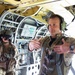 Two military personnel are photographed inside the cargo/crew compartment of a large military aircraft, likely a CH-47 Chinook or similar heavy-lift helicopter. The foreground figure, wearing a flight suit, multicam uniform, and aviation headset, appears to be gesturing with both arms extended outward. The interior shows exposed wiring, hydraulic lines, structural framing, and control panels painted in yellow-green primer.