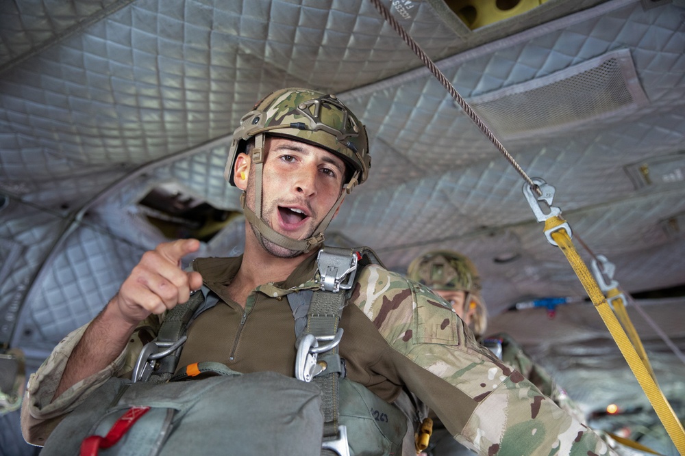 A military paratrooper is seated inside a military transport aircraft, wearing full jump gear including a camouflage helmet, parachute harness, and OCP/multicam uniform. He appears to be speaking or giving instructions while pointing toward the camera. A second soldier in similar gear is visible in the background.