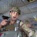A military paratrooper is seated inside a military transport aircraft, wearing full jump gear including a camouflage helmet, parachute harness, and OCP/multicam uniform. He appears to be speaking or giving instructions while pointing toward the camera. A second soldier in similar gear is visible in the background.