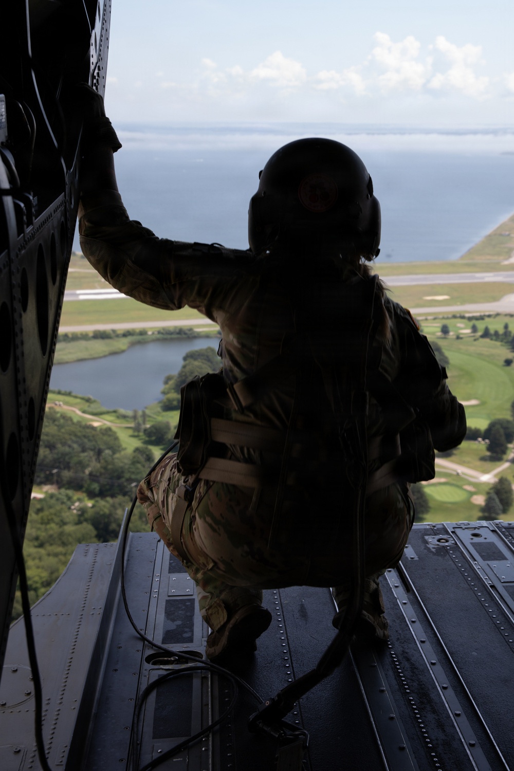 A military crew member in full flight gear and helmet crouches on the open rear ramp of a large helicopter, looking out over a coastal landscape below. The silhouetted figure is tethered by a safety cable and appears to be conducting aerial observation or monitoring a landing zone. The terrain below includes green fields, a small body of water, a runway or taxiway, and open water in the background.