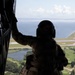 A military crew member in full flight gear and helmet crouches on the open rear ramp of a large helicopter, looking out over a coastal landscape below. The silhouetted figure is tethered by a safety cable and appears to be conducting aerial observation or monitoring a landing zone. The terrain below includes green fields, a small body of water, a runway or taxiway, and open water in the background.
