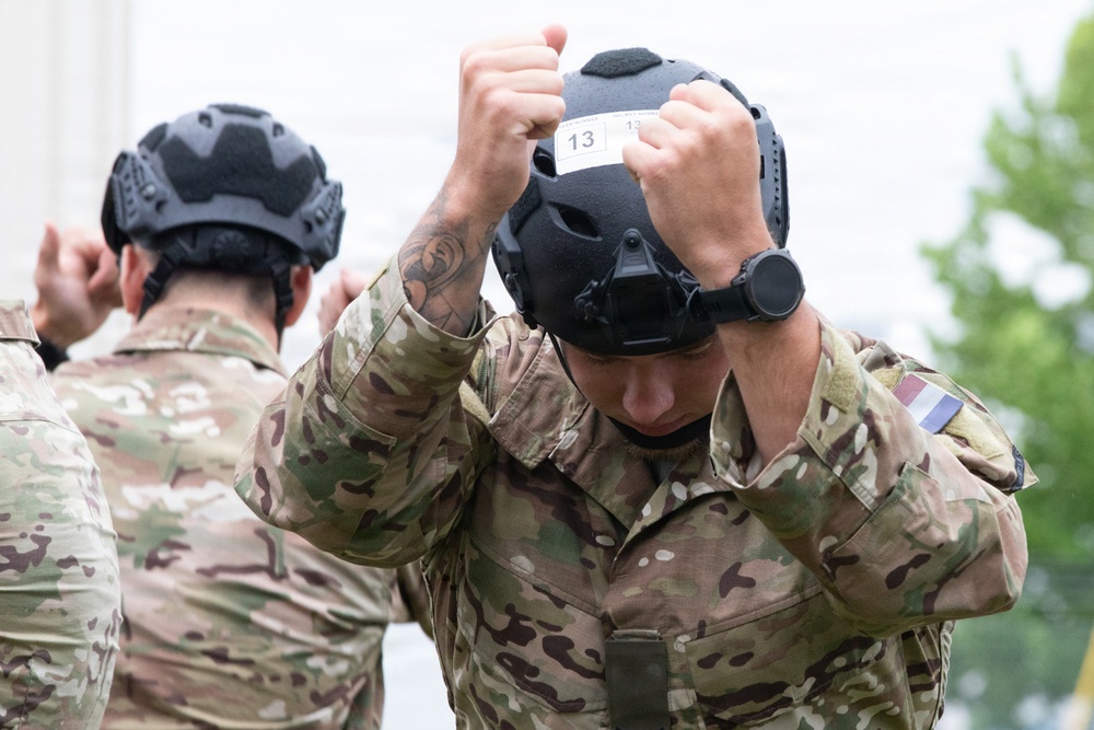 Two military personnel in MultiCam/OCP camouflage uniforms are donning tactical helmets. The foreground soldier is placing a black helmet labeled '13' onto his head, while a second soldier in the background performs the same action. The setting appears to be an outdoor area near a building with trees visible.
