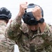 Two military personnel in MultiCam/OCP camouflage uniforms are donning tactical helmets. The foreground soldier is placing a black helmet labeled '13' onto his head, while a second soldier in the background performs the same action. The setting appears to be an outdoor area near a building with trees visible.