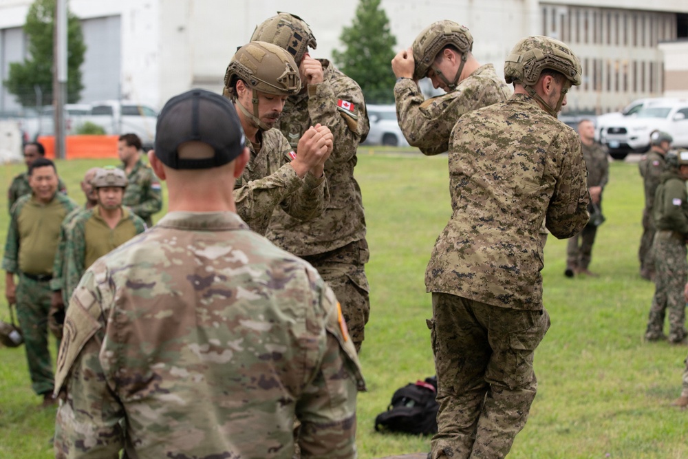 Military personnel from multiple nations are gathered on a grassy field, appearing to be preparing or adjusting equipment during a joint training exercise. Two soldiers wearing Canadian flag patches are prominently adjusting their tactical helmets in the foreground, while a soldier in US Army MultiCam uniform observes. Additional personnel in various camouflage patterns, including what appear to be Southeast Asian military uniforms, are visible in the background.