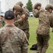Military personnel from multiple nations are gathered on a grassy field, appearing to be preparing or adjusting equipment during a joint training exercise. Two soldiers wearing Canadian flag patches are prominently adjusting their tactical helmets in the foreground, while a soldier in US Army MultiCam uniform observes. Additional personnel in various camouflage patterns, including what appear to be Southeast Asian military uniforms, are visible in the background.