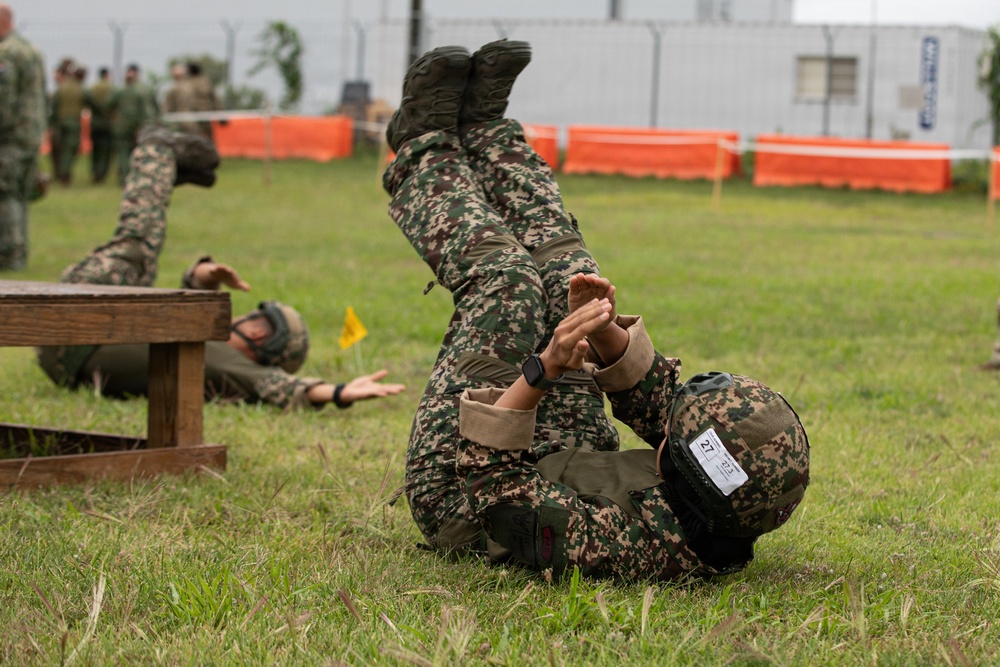 Military personnel in camouflage uniforms are engaged in a combat training exercise on a grass field. The foreground shows one soldier performing what appears to be a ground grappling or combatives technique, while another soldier is visible crawling under a wooden obstacle in the background. The setting appears to be an organized training course with orange barriers and a yellow flag marker visible.