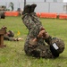 Military personnel in camouflage uniforms are engaged in a combat training exercise on a grass field. The foreground shows one soldier performing what appears to be a ground grappling or combatives technique, while another soldier is visible crawling under a wooden obstacle in the background. The setting appears to be an organized training course with orange barriers and a yellow flag marker visible.