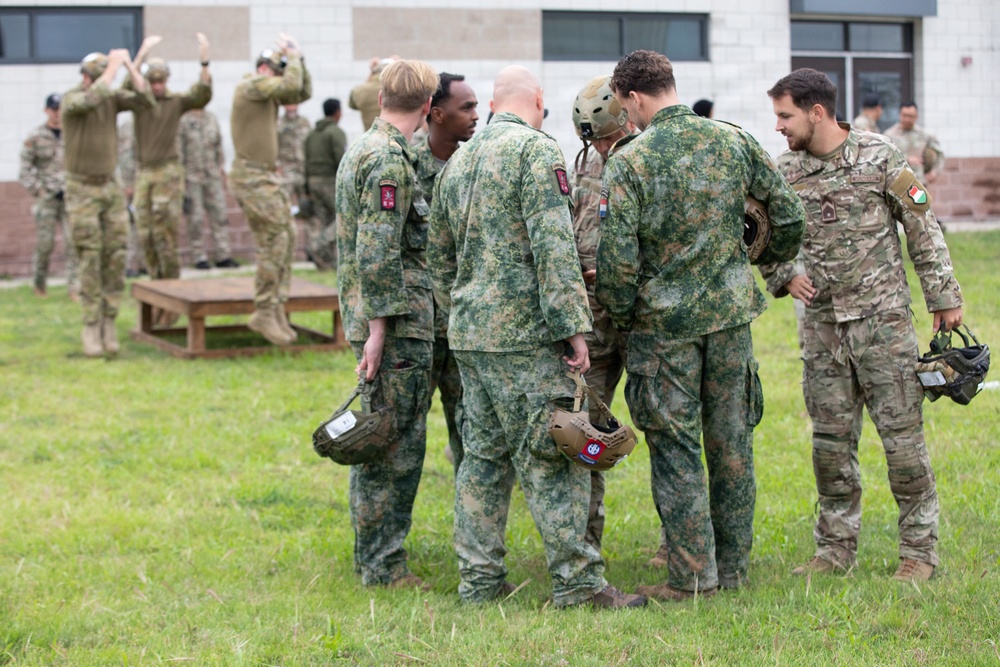 A group of military personnel in various camouflage uniforms are gathered outdoors on a grassy area near a building, appearing to be in conversation or a briefing huddle. Multiple nationalities appear to be represented based on the different uniform patterns and shoulder patches visible. In the background, additional soldiers are engaged in separate activities near what appears to be a cornhole board.