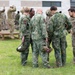 A group of military personnel in various camouflage uniforms are gathered outdoors on a grassy area near a building, appearing to be in conversation or a briefing huddle. Multiple nationalities appear to be represented based on the different uniform patterns and shoulder patches visible. In the background, additional soldiers are engaged in separate activities near what appears to be a cornhole board.