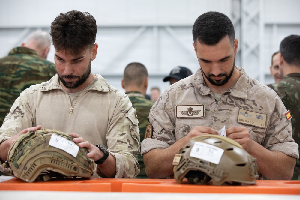 Two military personnel in desert/arid camouflage uniforms are examining tactical helmets placed on an orange surface, likely during an equipment inspection or issue event. The soldier on the right has a name tape reading 'DELGADO' and displays a Spanish flag patch along with wing insignia, indicating Spanish Armed Forces affiliation. Multiple other military personnel in various uniforms are visible in the background within what appears to be a tent or hangar facility.