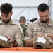 Two military personnel in desert/arid camouflage uniforms are examining tactical helmets placed on an orange surface, likely during an equipment inspection or issue event. The soldier on the right has a name tape reading 'DELGADO' and displays a Spanish flag patch along with wing insignia, indicating Spanish Armed Forces affiliation. Multiple other military personnel in various uniforms are visible in the background within what appears to be a tent or hangar facility.