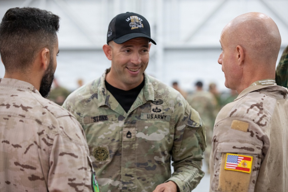 Three military personnel are engaged in conversation, with a U.S. Army soldier identified by name tape 'Stone' in the center wearing multicam uniform and a black cap. The soldier on the right wears a desert camouflage uniform with a combined U.S. and Spanish flag patch, indicating a joint or multinational military exercise. The setting appears to be a military facility or hangar with other personnel visible in the background.
