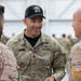 Three military personnel are engaged in conversation, with a U.S. Army soldier identified by name tape 'Stone' in the center wearing multicam uniform and a black cap. The soldier on the right wears a desert camouflage uniform with a combined U.S. and Spanish flag patch, indicating a joint or multinational military exercise. The setting appears to be a military facility or hangar with other personnel visible in the background.