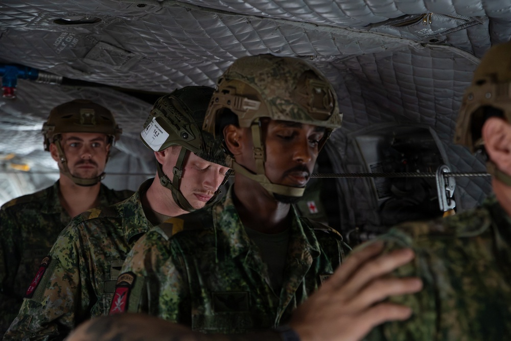Several military personnel in camouflage uniforms and tactical helmets are seated inside what appears to be a military aircraft or helicopter. At least one individual displays a red cross medical patch, suggesting the presence of a combat medic. The personnel appear to be in transit or preparing for an operation.