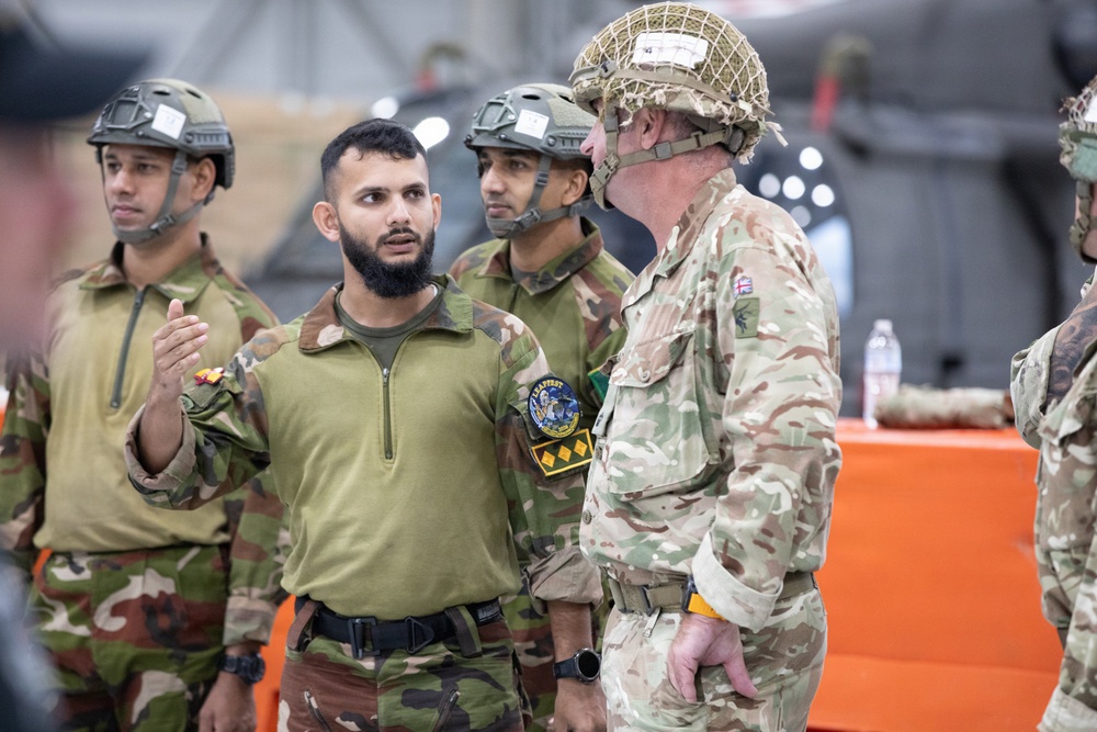 The image shows a group of military personnel from at least two different nations engaged in what appears to be a briefing or discussion inside a hangar or military facility. A soldier wearing an olive-green combat shirt with unit patches is speaking to a British Army soldier wearing MTP (Multi-Terrain Pattern) camouflage and a netted helmet. The interaction suggests a joint military exercise or training event between allied forces.