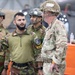 The image shows a group of military personnel from at least two different nations engaged in what appears to be a briefing or discussion inside a hangar or military facility. A soldier wearing an olive-green combat shirt with unit patches is speaking to a British Army soldier wearing MTP (Multi-Terrain Pattern) camouflage and a netted helmet. The interaction suggests a joint military exercise or training event between allied forces.