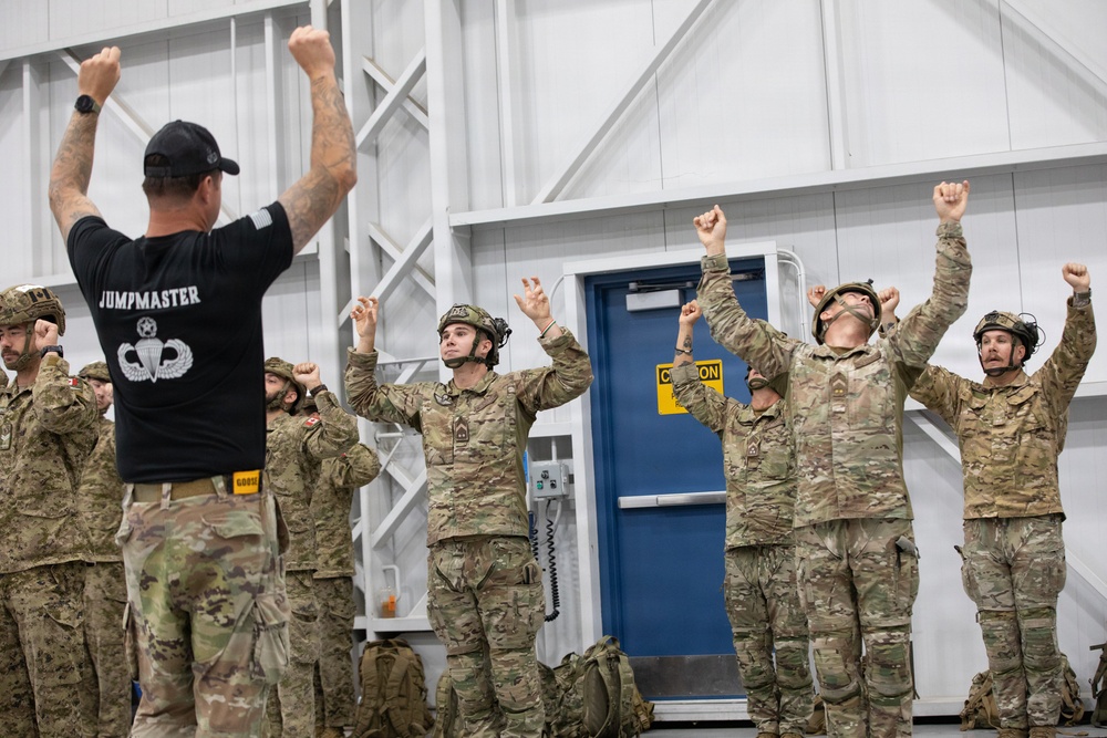A military jumpmaster instructor leads a group of soldiers in what appears to be pre-jump training or a parachute landing fall (PLF) drill inside a training facility. The instructor, wearing a black 'JUMPMASTER' t-shirt, faces the soldiers with arms raised while the soldiers in camouflage uniforms and helmets mirror the position. This appears to be airborne training, likely at a military jump school or airborne unit.