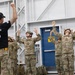 A military jumpmaster instructor leads a group of soldiers in what appears to be pre-jump training or a parachute landing fall (PLF) drill inside a training facility. The instructor, wearing a black 'JUMPMASTER' t-shirt, faces the soldiers with arms raised while the soldiers in camouflage uniforms and helmets mirror the position. This appears to be airborne training, likely at a military jump school or airborne unit.