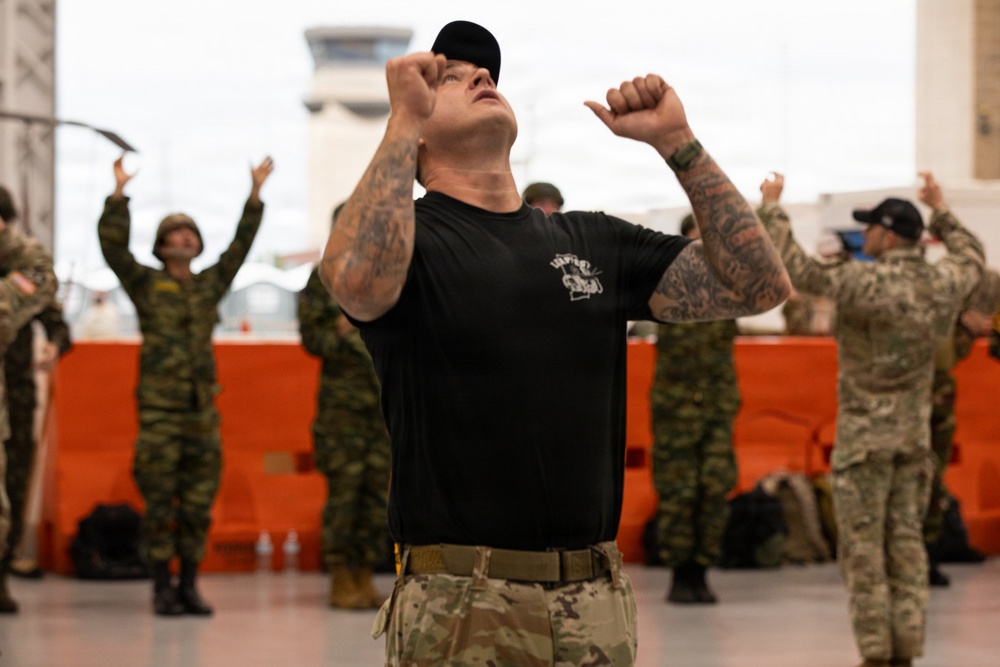 A heavily tattooed instructor or lead trainer in a black t-shirt and multicam pants leads a group of military personnel in a physical exercise or warm-up routine inside what appears to be a large hangar or terminal facility. The foreground figure has both fists raised upward with head tilted back, while soldiers in various camouflage uniforms mirror the movement in the background. The setting suggests a multinational military training event or exercise.