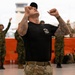 A heavily tattooed instructor or lead trainer in a black t-shirt and multicam pants leads a group of military personnel in a physical exercise or warm-up routine inside what appears to be a large hangar or terminal facility. The foreground figure has both fists raised upward with head tilted back, while soldiers in various camouflage uniforms mirror the movement in the background. The setting suggests a multinational military training event or exercise.