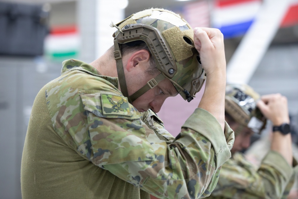 A military personnel member is adjusting a tactical helmet on their head in what appears to be an indoor facility. The soldier is wearing multicam/camouflage pattern uniform with a visible unit patch on the sleeve. A second similarly uniformed individual is visible in the background performing the same action.