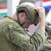 A military personnel member is adjusting a tactical helmet on their head in what appears to be an indoor facility. The soldier is wearing multicam/camouflage pattern uniform with a visible unit patch on the sleeve. A second similarly uniformed individual is visible in the background performing the same action.