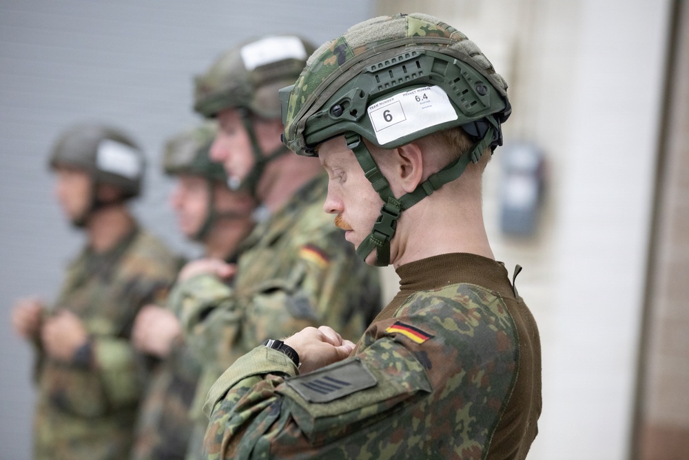 The image shows a line of military personnel wearing German Bundeswehr flecktarn camouflage uniforms and tactical helmets. The foreground soldier's helmet bears a label reading 'Team Number 6' and 'Helmet Number 6.4', suggesting a structured military exercise or competition. German flag patches are visible on the soldiers' sleeves, confirming their national affiliation.